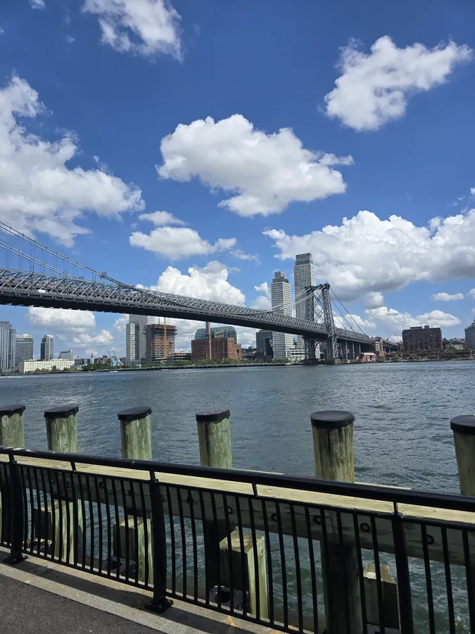 A bridge and city buildings seen from a waterfront railing under a bright sky