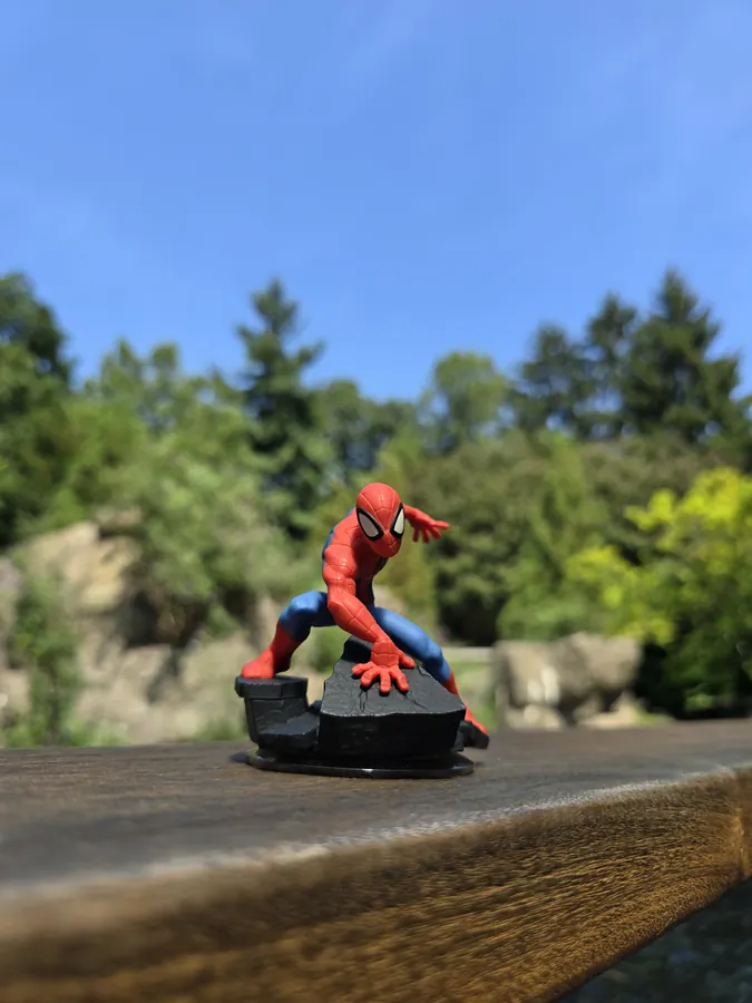 A small Spider-Man figure sitting on a wooden rail with trees behind it